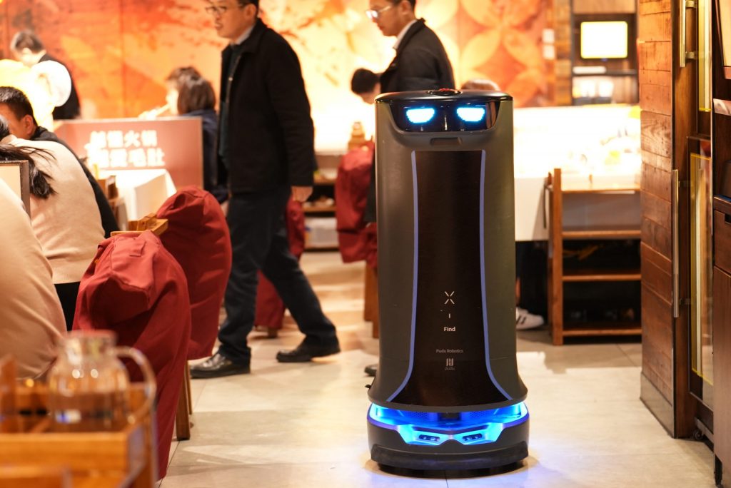 Ein schwarzer Robotik-Service-Roboter mit Blaulicht bewegt sich durch ein belebtes Restaurant mit sitzenden und gehenden Menschen.