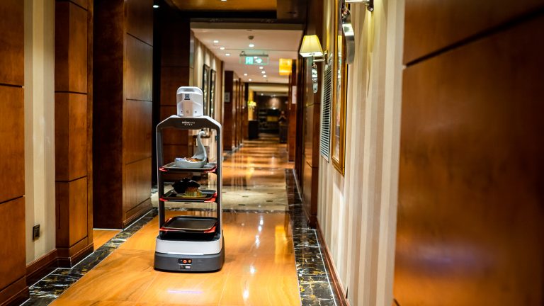 Ein Serviceroboter navigiert durch autonome Navigation durch polierte Böden und Holzwände in einem Hotelflur und liefert mit fortschrittlicher Robotik-Technologie effizient Gegenstände auf Tabletts aus.