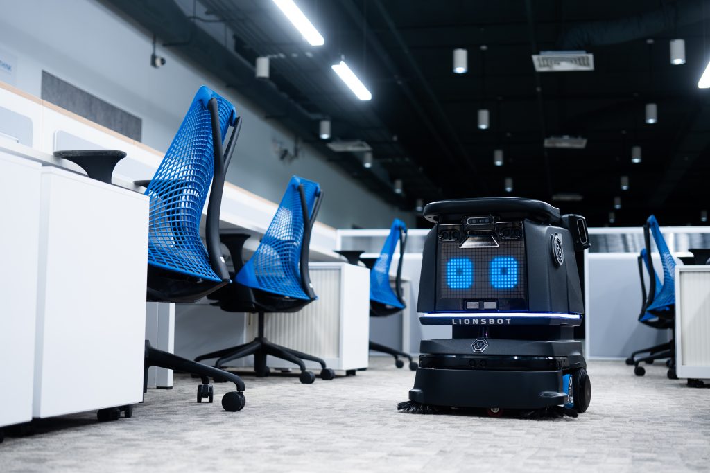 Ein schwarzer Putzroboter bewegt sich zwischen blauen Bürostühlen in einem modernen, leeren Office Space.