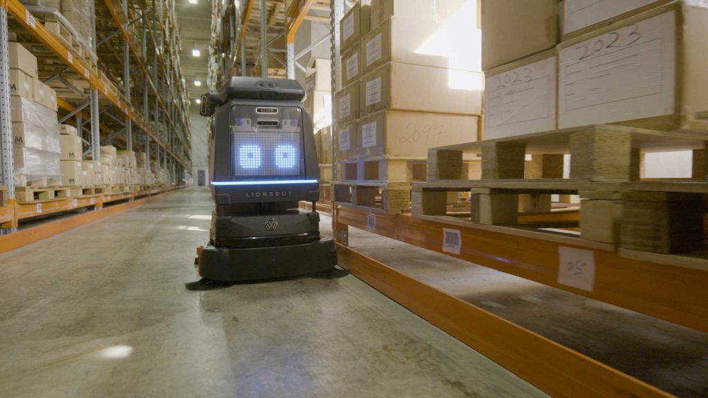 Ein kleiner Robotik Roboter mit digitalen Augen bewegt sich entlang eines Ganges in einem Lager mit Regalen und Paletten.