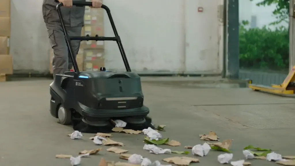Eine Person benutzt einen Putzroboter, um den Boden einer Lagerhalle von Papier und Laub zu reinigen.