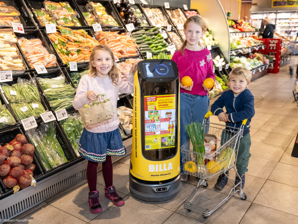 Drei lächelnde Kinder mit Lebensmitteln stehen neben einem Kettybot in der Lebensmittelabteilung eines Supermarkts.