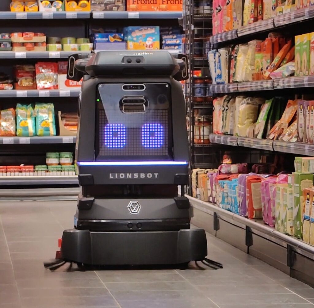 Ein Reinigungsroboter von Salesmotion Robotics mit digitalen Augen arbeitet in einem Supermarktgang zwischen den Regalen mit Lebensmitteln.