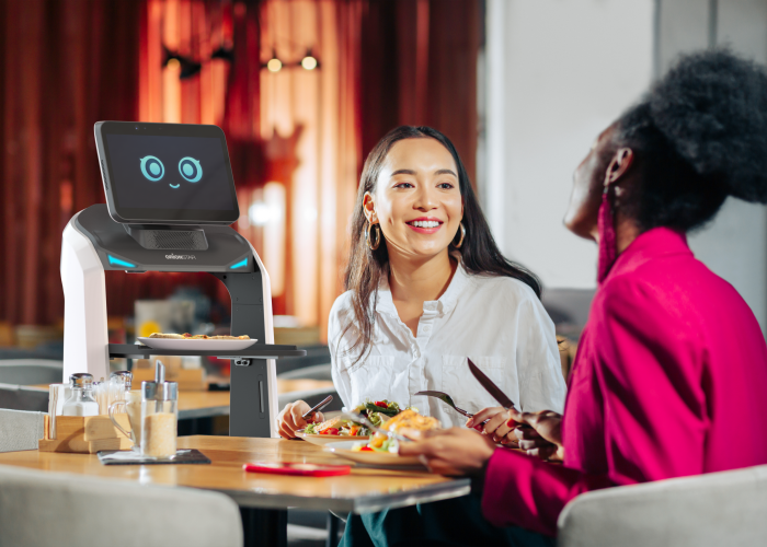 Zwei Frauen, die in einem Restaurant speisen, werden von einem freundlichen Gastronomie Roboter bedient, der ein lächelndes digitales Gesicht auf seinem Bildschirm zeigt.