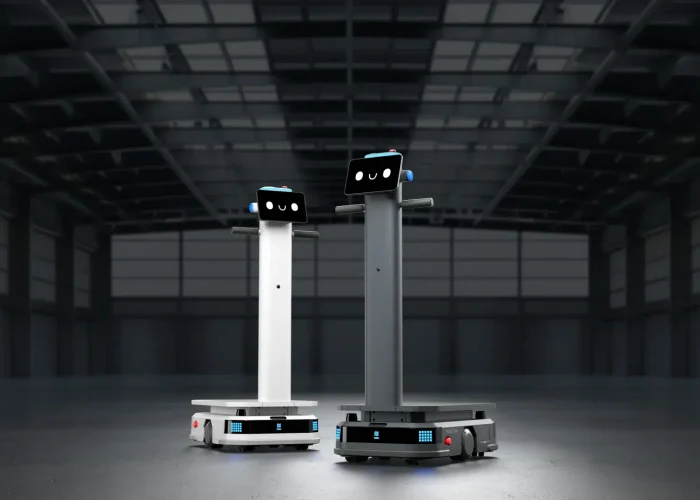 Zwei große autonome Lieferroboter mit Bildschirmgesichtern stehen in einer leeren Industriehalle mit hohen Decken und zeigen die Zukunft der Robotik-Innovation.